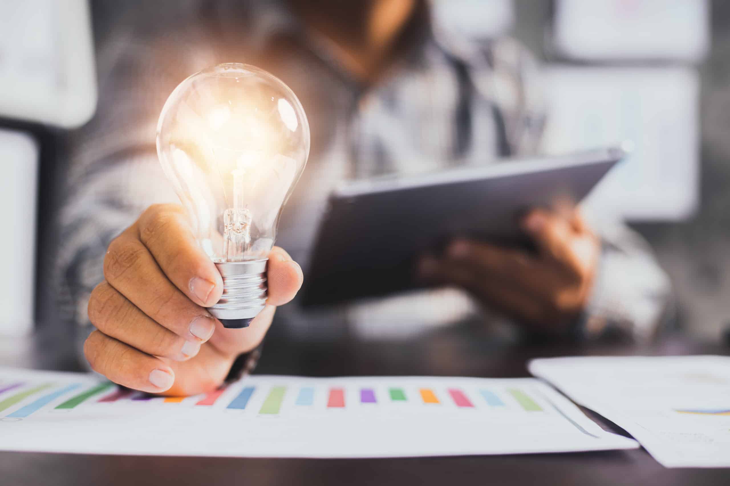 Successful business idea and creative innovation concept, close up business man holding lighting bulb and tablet with financial graph on table